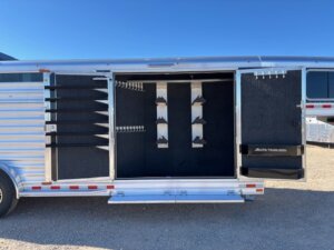 An Elite 4+1 Combo horse trailer with its side door open reveals a spacious Side Tack area with saddle racks, bridle hooks, and shelves, all lined with dark material. The trailer is parked on gravel under a clear blue sky. -Stillwater Trailer Sales
