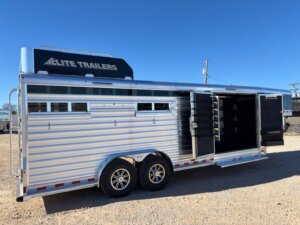A silver Elite 4+1 Combo livestock trailer with &ldquo;Elite Trailers&rdquo; branding is parked on gravel. The Side Tack compartment and storage areas are open, revealing shelves inside. Dual axles with alloy wheels rest under a clear blue sky. -Stillwater Trailer Sales