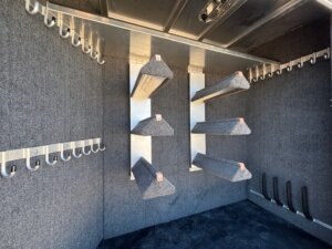 Interior of a Side Tack room with carpeted saddle racks mounted on the wall, rows of metal bridle hooks along two walls, and several vertical slots near the floor for holding saddle pads or other Elite 4+1 Combo equipment. -Stillwater Trailer Sales