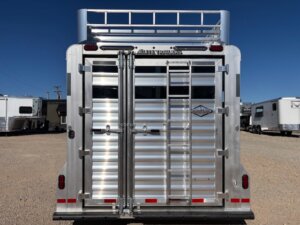 A rear view of an Elite 4+1 Combo silver aluminum livestock trailer with vertical and horizontal bars, two doors, tail lights, Side Tack storage, and a ladder leading to a roof rack. The trailer is parked on a gravel lot under a clear blue sky. -Stillwater Trailer Sales