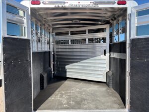 Interior view of an empty, open Elite 4+1 Combo livestock trailer with metal walls, rear gate, side tack, non-slip flooring, and ventilated panels. The trailer is clean and well-lit by sunlight. -Stillwater Trailer Sales