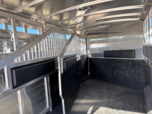 Interior of an empty Elite 4+1 Combo livestock trailer with metal walls, partitions, ventilation slats, padded side panels, and a non-slip black floor. Sunlight filters in through windows and gaps in this Elite trailer's well-ventilated design. -Stillwater Trailer Sales