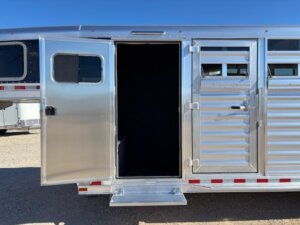 A metallic Elite 4+1 Combo livestock trailer with a Side Tack, open side door, and fold-down step reveals a dark interior. The trailer features horizontal slats and a locked gate beside the open door against a clear blue sky. -Stillwater Trailer Sales