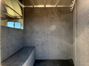 A small, carpeted Side Tack interior with gray walls, a built-in bench on the left, metal ceiling, window on the left wall, and ceiling hooks on the right. Sunlight shines through the window in this Elite 4+1 Combo space. -Stillwater Trailer Sales