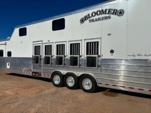 A large white and silver Bloomer horse trailer with multiple windows and vents, parked on a dirt surface under a clear blue sky. -Stillwater Trailer Sales