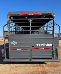 Rear view of a black Swift Built Tack Box Combo livestock trailer with metal bars and red reflective tape, parked on a dirt lot under a clear blue sky. The Swift Built logo and another emblem are visible on the back. -Stillwater Trailer Sales