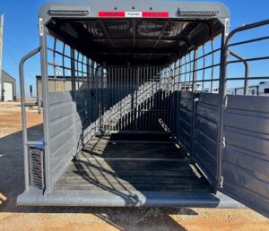 The image shows the open rear view of an empty Swift Built livestock trailer with metal sides and a grated interior gate, parked on a gravel lot under a clear blue sky. This 22' model features a practical Tack Box Combo for added convenience. -Stillwater Trailer Sales