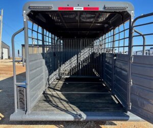 A photo of the empty interior of a gray metal Swift Built livestock trailer, featuring grated sides, a tack box, and a gate at the far end, parked outdoors on gravel under a clear blue sky. -Stillwater Trailer Sales