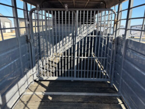 A Swift Built 22' metal livestock trailer with a Tack Box Combo features an open gate and barred partitions, allowing sunlight to cast shadows on the wooden floor. The trailer is stationary and empty. -Stillwater Trailer Sales