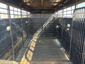Interior of an empty livestock trailer with metal walls and a wooden floor, sunlight casting shadows through the bars and open sections, creating a pattern on the floor. -Stillwater Trailer Sales