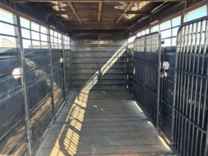 The image shows the interior of an empty livestock trailer with metal walls, slatted side panels, gated partitions, and a wooden floor, all viewed from the rear with light streaming in. -Stillwater Trailer Sales