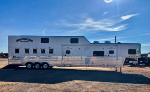 A large silver Bloomer horse trailer with multiple windows and three axles is parked on a gravel lot under a clear blue sky. -Stillwater Trailer Sales