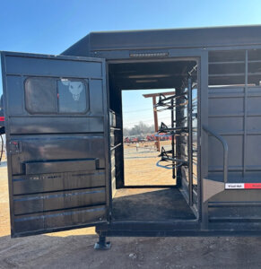 A black livestock trailer with its side door open, revealing metal interior bars and racks. The ground is dirt, and some equipment and fencing are visible in the background under a clear blue sky. -Stillwater Trailer Sales