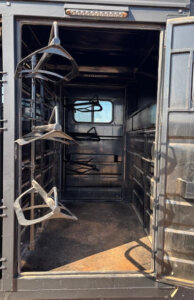 Open door of a black metal trailer showing an interior with saddle racks mounted on the left wall and a window in the back, with sunlight streaming onto the floor. -Stillwater Trailer Sales