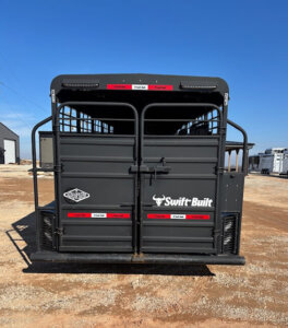 A black livestock trailer with Swift Built branding is parked on a gravel lot under a clear blue sky, with other trailers and a building visible in the background. -Stillwater Trailer Sales