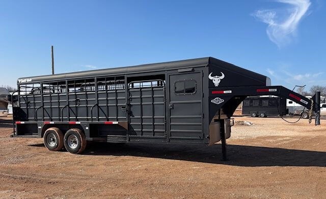 A large black livestock trailer with a gooseneck hitch is parked on a dirt lot under a sunny sky. The trailer has multiple side openings, two axles, and a white bull head logo on the front. -Stillwater Trailer Sales