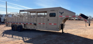 A beige gooseneck livestock trailer with dual axles stands on a dirt lot under a clear sky. The trailer has open side slats, red-and-white reflective tape, and a bull head logo on the front. -Stillwater Trailer Sales