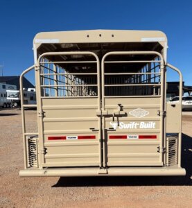 A tan Swift Built 28' Stock Trailer is parked on gravel, seen from the rear, with metal bars, latches, and red reflectors. Other trailers and buildings are visible in the background under a clear blue sky. -Stillwater Trailer Sales