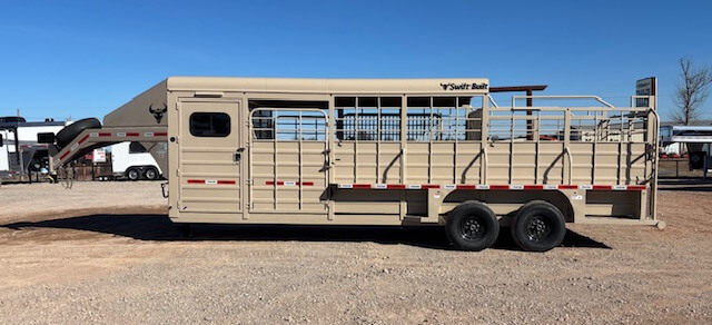 A tan livestock trailer with two axles, side rails, and a gooseneck hitch is parked on a gravel lot under a clear blue sky. The trailer has Swift Built branding on the upper side. -Stillwater Trailer Sales