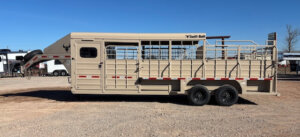 A tan gooseneck livestock trailer with metal bars and double axles is parked on a gravel lot under a clear blue sky. The trailer has Swift Built branding and is designed for transporting animals. -Stillwater Trailer Sales