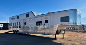 A large white gooseneck horse trailer with living quarters, multiple windows, and three axles is parked on a dirt lot under a clear blue sky. -Stillwater Trailer Sales