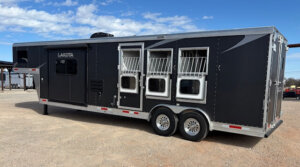 A large black and silver horse trailer with three barred windows, dual axles, and the brand name Lakota visible on the side, parked on gravel under a blue sky with scattered clouds. -Stillwater Trailer Sales