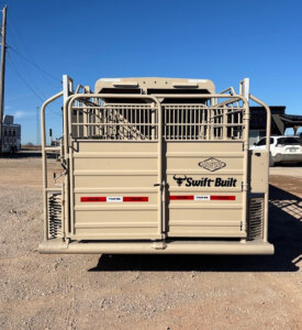 A beige livestock trailer with the Swift Built logo is parked on a gravel lot under a clear blue sky, viewed from the rear with its gate closed. -Stillwater Trailer Sales