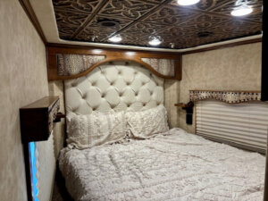 A cozy bedroom with a tufted white headboard, intricate ceiling tiles, soft beige walls, and a bed covered with a textured white bedspread. Wooden accents and a window with a woven blind complete the space. -Stillwater Trailer Sales