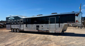 A large silver and black Bloomer gooseneck livestock trailer with multiple windows and three axles is parked on a gravel lot under a clear blue sky. -Stillwater Trailer Sales