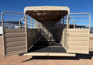 Open 28' Swift Built stock trailer with both rear doors wide open, revealing an empty interior with metal railings and a black rubber floor mat. The trailer is parked on gravel under a clear blue sky. -Stillwater Trailer Sales