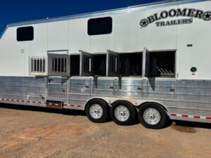 A large white Bloomer horse trailer with three axles is parked on dirt. Several stall doors and windows are open, revealing the interior designed for transporting horses. The trailer is clean and has a shiny metallic lower section. -Stillwater Trailer Sales