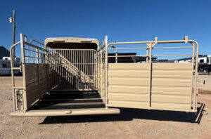 A beige livestock trailer with its rear gate open, showing a ramp and interior metal bars, is parked on a gravel surface under a clear blue sky. -Stillwater Trailer Sales