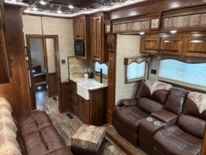 A cozy RV interior with brown leather sofas, a farmhouse sink, wooden cabinets, overhead storage, patterned ottoman, and wood-style flooring, illuminated by recessed ceiling lights. A door at the back leads to another room. -Stillwater Trailer Sales