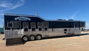 A large, shiny silver and black Bloomers horse trailer with multiple windows and vents is parked on a dirt lot under a clear blue sky. -Stillwater Trailer Sales