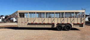 A beige Swift Built 28' stock trailer with open slats and dual axles is parked on a gravel lot under a clear blue sky. -Stillwater Trailer Sales