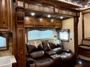 Luxurious RV interior with dark brown leather reclining seats, ornate wood cabinetry and trim, decorative ceiling panels, window shades, and carpeted steps leading up to another section. A small sink is visible on the left. -Stillwater Trailer Sales