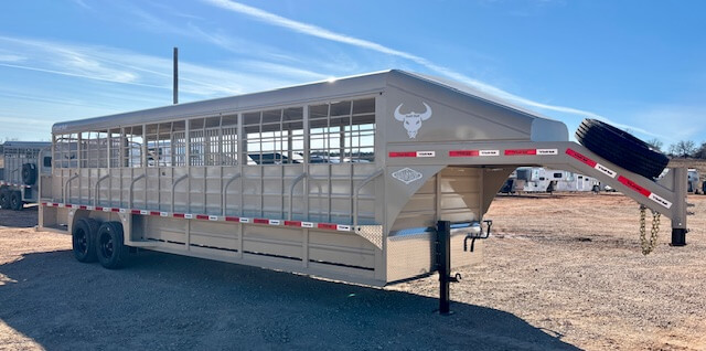 A large tan Swift Built 28' Stock Trailer with open sides, dual rear axles, and a gooseneck hitch is parked on a gravel lot under a clear blue sky. A bull head logo is visible on the front of this stock trailer. -Stillwater Trailer Sales