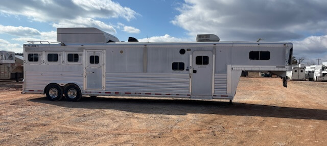 A large white gooseneck horse trailer with multiple windows and two axles is parked on a dirt lot under a partly cloudy sky. -Stillwater Trailer Sales