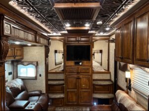 Luxurious RV interior with ornate wood cabinets, patterned ceiling, leather seating, wall-mounted TV, and small windows with shades. Two twin beds with pillows are built into the back wall on either side of the TV. -Stillwater Trailer Sales