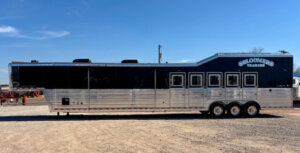 A large silver and black gooseneck trailer with the words BLOOMERS TRAILERS on the side, parked on a dirt lot under a clear blue sky. The trailer has six windows and three axles. -Stillwater Trailer Sales