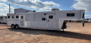A large silver gooseneck horse trailer with living quarters is parked on a dirt lot under a partly cloudy sky. The trailer has multiple windows, dual axles, and rooftop air conditioning units. -Stillwater Trailer Sales