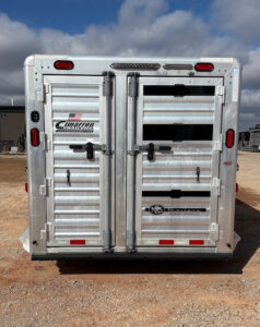 The image shows the closed rear doors of a silver Cimarron Stock Combo livestock trailer parked on gravel, with visible company logos, red and white reflectors, and latches securing the 26' doors. The sky is partly cloudy in the background. -Stillwater Trailer Sales