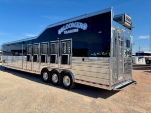 A large, shiny horse trailer with six windows and vents on the side, labeled Bloomer Trailers, sits parked on gravel under a clear blue sky. -Stillwater Trailer Sales