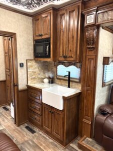 A cozy RV kitchen with wood cabinets, a farmhouse sink, black faucet, stone backsplash, microwave, and decorative accents. Brown leather seating and wood flooring complete the warm, rustic look. -Stillwater Trailer Sales