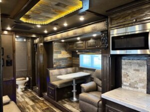 Interior of a luxury RV featuring a dining booth, leather seating, wood accents, overhead cabinets, a modern microwave, and a view into a small bathroom with a toilet. Warm lighting highlights the upscale finishes. -Stillwater Trailer Sales