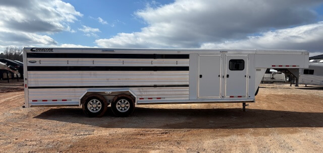 A large silver Cimarron 26' Stock Combo livestock trailer with black accents and two axles is parked on gravel under a partly cloudy sky. -Stillwater Trailer Sales