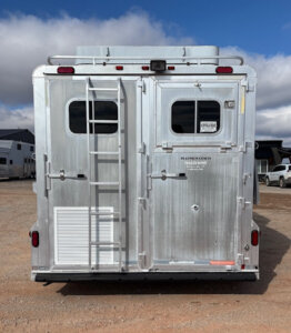 The rear view of a silver trailer with two doors, a ladder on the left door, a vent, windows, and various stickers. The trailer is parked on gravel with other vehicles and buildings in the background under a partly cloudy sky. -Stillwater Trailer Sales