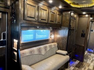 A cozy RV interior featuring wooden cabinets, a beige cushioned sofa with a patterned cover, small pillows, overhead lighting, and wood-style flooring. A window with blinds lets in natural light beside the sofa. -Stillwater Trailer Sales