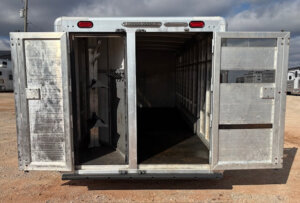 A 26' Cimarron stock combo trailer stands on a dirt lot under a partly cloudy sky, both rear doors open to reveal a dark interior and saddle racks lining the left side. -Stillwater Trailer Sales