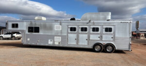 A large silver gooseneck horse trailer with multiple windows and compartments is parked on gravel under a partly cloudy sky. -Stillwater Trailer Sales